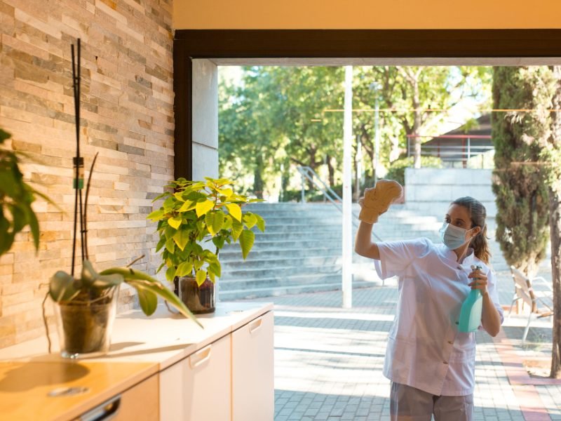 Front view of young woman in uniform and wearing a face mask cleaning windows of spa center from outside.